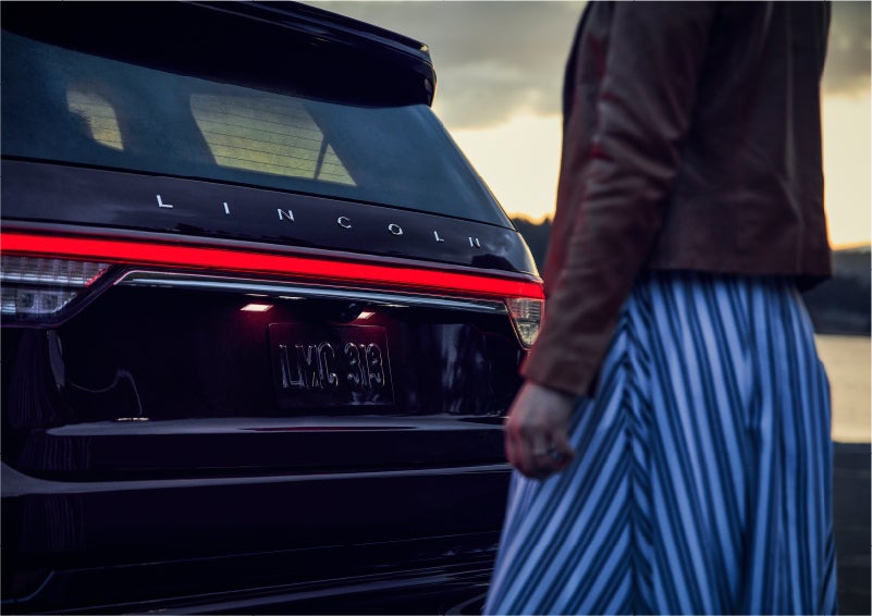 A person is shown near the rear of a 2023 Lincoln Aviator® SUV as the Lincoln Embrace illuminates the rear lights | Sheehy Lincoln of Gaithersburg in Gaithersburg MD